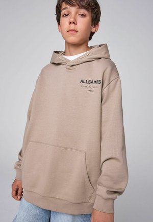 Ung person iført beige ALLSAINTS hættetrøje med forlomme, stående mod en ensfarvet lys grå baggrund.