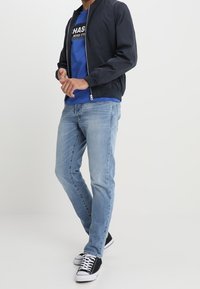 Chaqueta bomber navy con cremallera, camiseta azul con estampado gráfico, Jeans de mezclilla azul claro, zapatillas en blanco y negro, de pie sobre un fondo neutro.