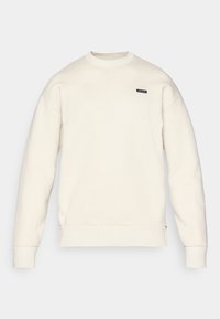 Sweatshirt beige clair avec un col rond, des bords-côtes au niveau des poignets et de l'ourlet, et un petit logo noir sur la poitrine. Texture douce et coupe décontractée.