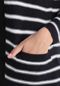 Granatowo-biały dzianinowy kardigan w paski z kieszenią, ściągaczami na rękawach i luźnym krojem. Tkanina ma fakturowany wygląd.