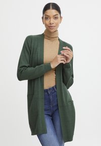 Cardigan verde intrecciato con apertura frontale, maniche lunghe e due tasche anteriori, abbinato a un dolcevita beige e jeans azzurri.