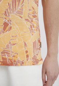 Camisa de manga corta en naranja y amarillo con un estampado de hojas. Fabricada con una tela suave, tiene un dobladillo recto y una pequeña etiqueta de la marca en el costado.