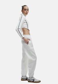 Witte cropped pullover met zwarte zijstrepen, gecombineerd met witte joggers met zakken. Het model draagt witte sneakers met zwarte accenten.