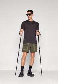 T-shirt grigio scuro a maniche corte con logo, pantaloni corti verde oliva con tasche frontali, sneakers nere e bastoni da trekking.