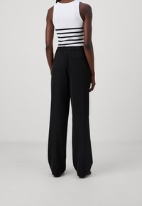 Nainen, jolla on päällä hihaton valkoinen paita mustilla raidoilla ja väljät mustat leveälahkeiset housut, seisoo selin yksinkertaista taustaa vasten.