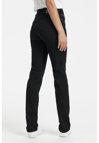 Persona vestida con jeans ajustados negros y zapatillas blancas, de pie con una mano relajada a su lado.