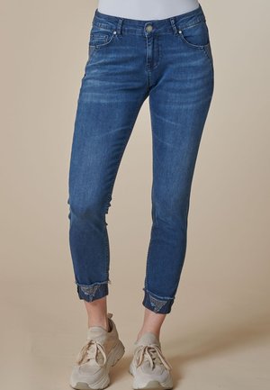 Blaue enge Denim-Jeans mit ausgefransten, hochgekrempelten Bündchen, getragen mit beigen klobigen Sneakern auf beige Hintergrund.