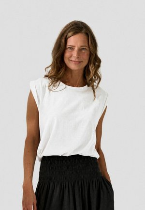 Femme aux cheveux bruns ondulés portant un haut blanc sans manches et une jupe noire, debout devant un fond gris clair uni.