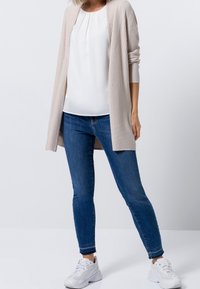 Cardigan a coste beige sopra una camicetta bianca, abbinato a jeans skinny blu e scarpe da ginnastica bianche. Il cardigan presenta una chiusura aperta e maniche lunghe.