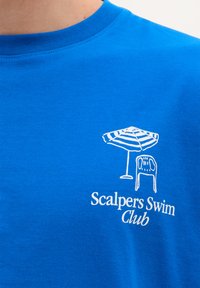 Blå bomulds T-shirt med et hvidt grafik af en strandparasols og stol, med teksten "Scalpers Swim Club" nedenfor. Korte ærmer, rund halsudskæring.