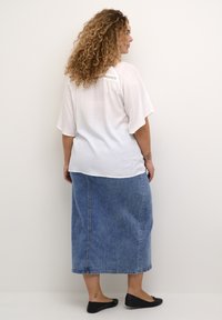 Hvid kortærmet top med en blondeaccent på ryggen, parret med en lang blå denimnederdel og sorte flade sko. Glat tekstur, casual design.