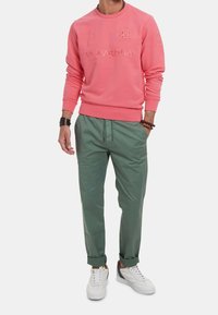 Růžová mikina s kulatým výstřihem a žebrovanými lemy, s vyraženým logem a číslem. Zelené kalhoty se šňůrkou na stažení a bílé tenisky.