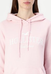 Detailní záběr osoby v světle růžové mikině s kapucí, na které je vyšitý nápis "Hollister California" a logo ptáka na přední straně.