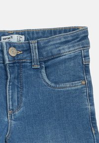 Jeans iz džinsa s srednje modrim odtenkom, z zapenjanjem na gumb, petimi žepi in kontrastnim šivom ob robovih. Notranja etiketa z blagovno znamko je vidna na pasu.