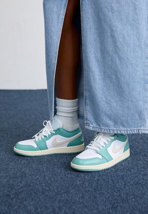 Pieds portant des baskets Nike turquoise et blanches avec des chaussettes blanches, partiellement couverts par une longue jupe en jean bleu clair, debout sur un tapis bleu.
