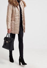 Giacca trapuntata beige con cappuccio foderato in pelliccia, abbinata a pantaloni neri aderenti, borsa nera e stivali neri con tacco alla caviglia.