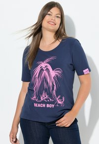 Usmívající se žena s dlouhými hnědými vlasy, oblečená v námořnickém modrém tričku s růžovou grafikou dlouhosrstého psa, kraba a textem "BEACH BOY".