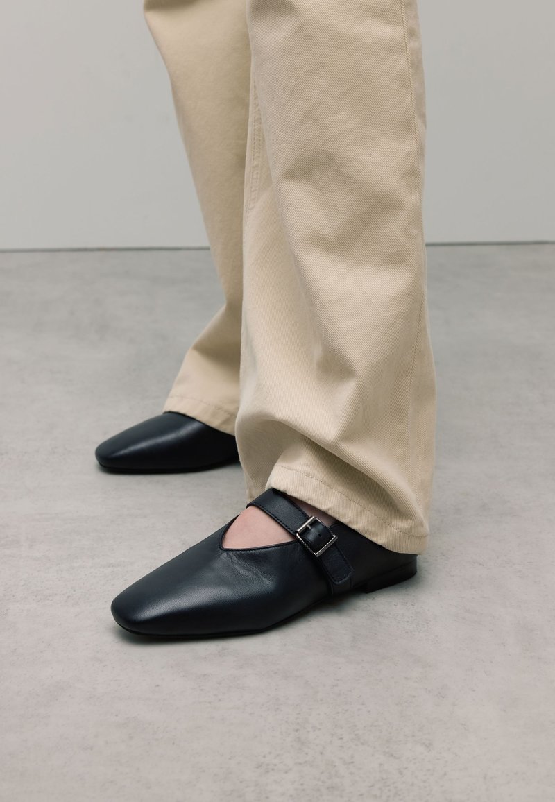 Zwarte leren schoenen met een spitse neus, voorzien van een uitsnede en een gespsluiting aan de zijkant, gecombineerd met lichtbeige broeken.