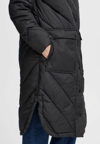 Černý péřový kabát s designem v stoupající šev, patentkami, velkou kapsou a bočními rozparky pro snadnější pohyb.