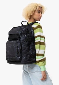 Svart och grå ryggsäck med mönster, flera fickor och nätdetaljer, bärs på ryggen, tillsammans med en flerfärgad randig tröja.