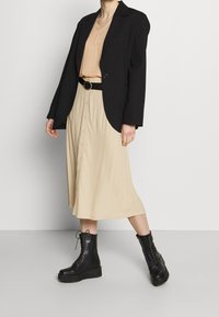 Blazer noir sur un haut beige et une jupe boutonnée couleur crème, associé à des bottes de combat à lacets noires. Texture lisse, design décontracté.