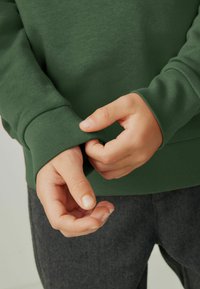 Felpa verde con polsini a coste, mostrante mani che regolano la manica. Il tessuto appare liscio senza motivi visibili, abbinato a pantaloni grigi scuri.