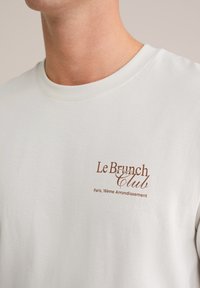 Balta kokvilnas T-kreklu ar īsām piedurknēm, apaļu kaklu un brūna teksta logo "Le Brunch Club, Parīze, 16. rajons" uz krūšu.
