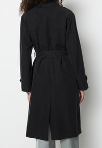Černý trenčkot s páskem, hladkou texturou, dlouhými rukávy a zadním rozparkem. Má klasický límec a nastavitelné manžetové pásky.