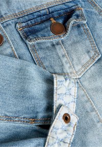 Helle blaue Denim-Stoff mit kontrastierender brauner Naht. Verfügt über eine Fronttasche mit einem Metallknopf und einem blumigen Innenfutter.