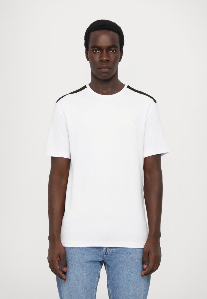 White short-sleeve t-shirt with black shoulder accents and a small gold logo on the chest. Made of smooth cotton fabric. Simple design.