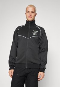 Mustvalge tõmblukuga jakk kõrge kaelusega, millel on valged äärised, broderitud logo ning soonikkoes varrukate ja hem. Sile tekstuur.