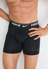 Schwarze Nike Boxershorts mit elastischem Bund, auf dem das Markenlogo in Weiß und einem orangefarbenen Akzent zu sehen ist. Weiches, anliegendes Material mit glatter Textur.