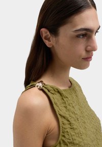 Olivgrön strukturerad topp med en rund metallisk detalj på axeln. Har en avslappnad passform och en modern design.