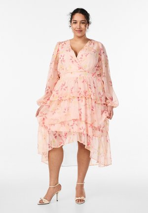 Kobieta ubrana w warstwową, brzoskwiniowo-różową sukienkę w kwiaty z długimi przezroczystymi rękawami i asymetrycznym dołem, w połączeniu z kremowymi szpilkami na kostkę z paskiem.