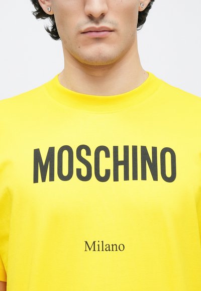 Sárga pamut póló, vastag fekete "MOSCHINO" felirattal a mellkason, alatta kisebb betűkkel "Milano". Klasszikus kerek nyakkivágás.