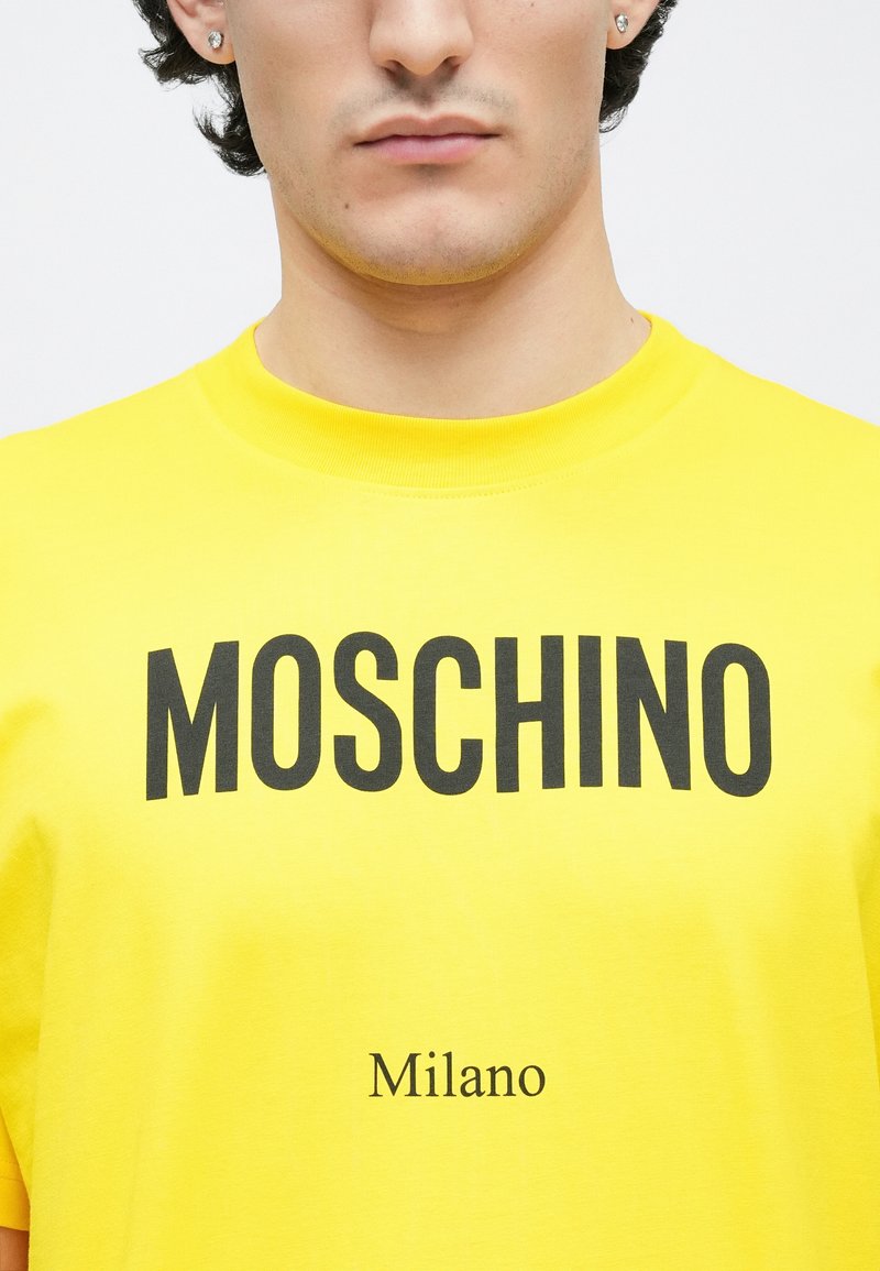 Gul bomuld T-shirt med fed sort tekst "MOSCHINO" tværs over brystet og "Milano" nedenunder i mindre skrift. Klassisk rund halsudskæring.
