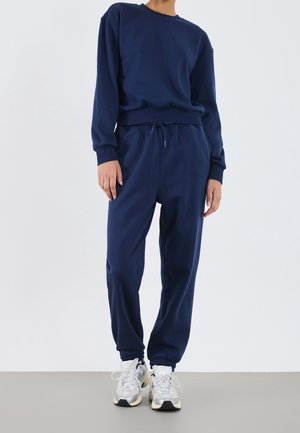 Persona con sudadera azul marino y pantalones deportivos a juego, con zapatillas deportivas blancas, de pie sobre un suelo gris y fondo blanco.