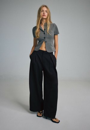 Young woman stands with hands in pockets wearing short-sleeve gray blazer, wide-leg black trousers, and black heeled sandals against plain background.