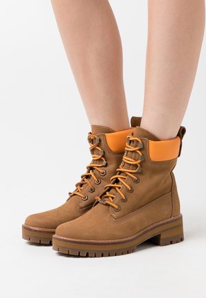 Bottes en cuir marron avec des lacets et un col orange. Elles présentent des œillets en métal, une semelle robuste et des détails de couture sur les côtés.