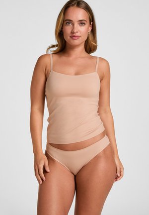 Woman with shoulder-length blonde hair wearing nude-colored camisole and matching underwear, standing against a plain light background.