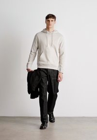 Harmaa-beige huppari, jossa on etutasku, yhdistettynä tummiin kuviollisiin housuihin. Malli tuo mustaa takkia, kävellen neutraalilla taustalla.