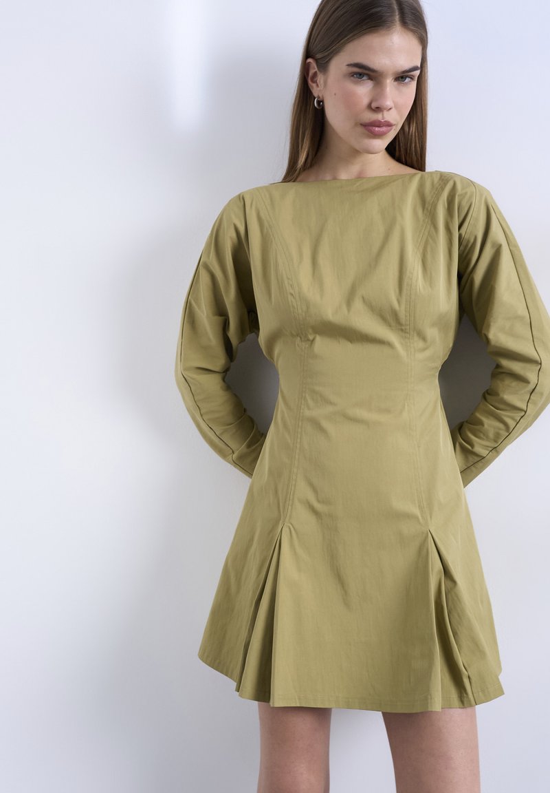 Νεαρή γυναίκα με ίσια μαλλιά, φορώντας ένα μακρυμάνικο ελιάς πράσινο φόρεμα με πιέτες, στέκεται μπροστά σε έναν απλό φωτεινό τοίχο.