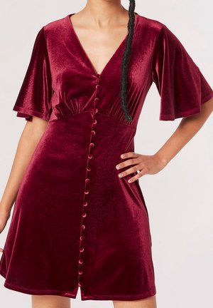 Kobieta w głęboko czerwonej aksamitnej sukience z falbanowymi rękawami i guziczkami obciągniętymi tkaniną z przodu, z ręką na biodrze, na jednolitym tle.