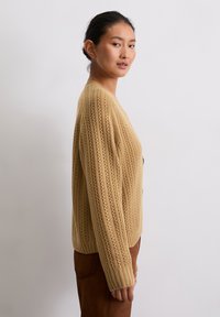 Strickjacke in einem hellen Beige mit gerippter Textur und Durchbrüchen. Sie hat lange Ärmel und eine Knopfverschluss vorne.
