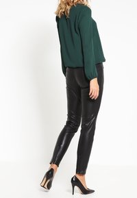 Blusa verde a maniche lunghe abbinata a pantaloni in pelle nera aderenti e scarpe con tacco alto nere, con dettagli con zip alle caviglie.