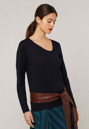 Vrouw met donker haar in een paardenstaart, gekleed in een zwarte asymmetrische top, brede bruine leren riem en een blauwgroene gestreepte rok, kijkt naar beneden.