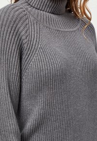 Pull à col roulé en maille côtelée grise avec des détails de coutures diagonales sur la poitrine et les épaules, porté par une personne avec des mèches de cheveux châtain clair visibles.