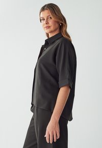 Camicia nera a maniche corte, con bottoni e vestibilità comoda. Tessuto liscio, colletto a rever e orlo arrotondato. Abbinata a pantaloni coordinati.