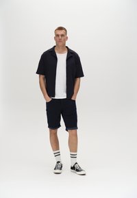 Kurzarm-Hemden in Marineblau über einem weißen T-Shirt, kombiniert mit marineblauen Shorts. Weiße Socken mit schwarzen Streifen und schwarze Turnschuhe runden den Look ab.