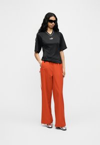 Γυναίκα που φοράει μαύρο πουκάμισο Puma, πορτοκαλί παντελόνια με φαρδύ πόδι, ασημί παπούτσια και μαύρα γυαλιά ηλίου, στέκεται με το ένα χέρι στην τσέπη μπροστά σε λευκό φόντο.